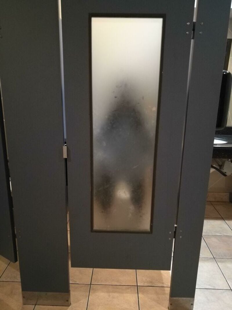 Frosted bathroom stall door with a blurry figure behind it.