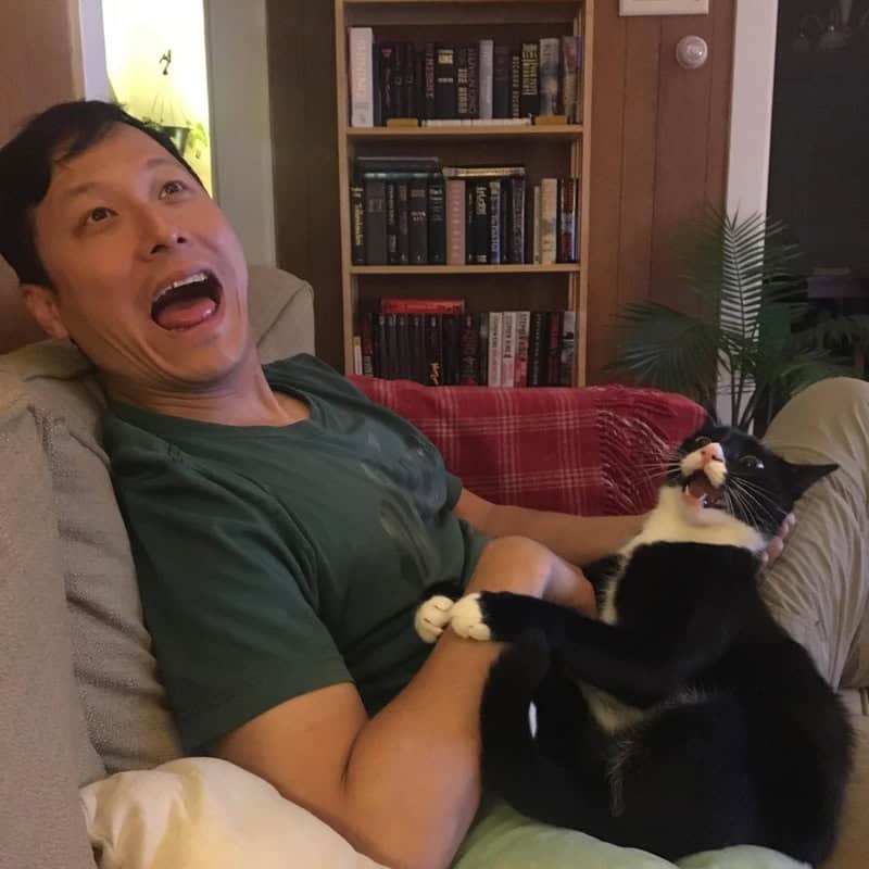 Man with black hair and green shirt playing with a black and white cat on a beige couch.