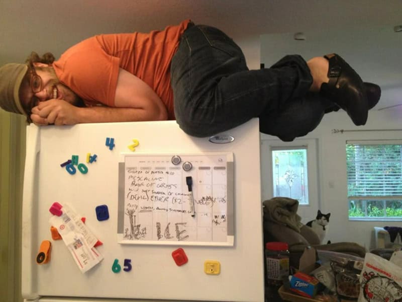 A woman lying sideways on top of a refrigerator, with her head resting on her hand.
