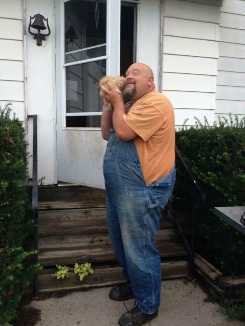 Man hugging a small dog outside a house with stairs and bushes.