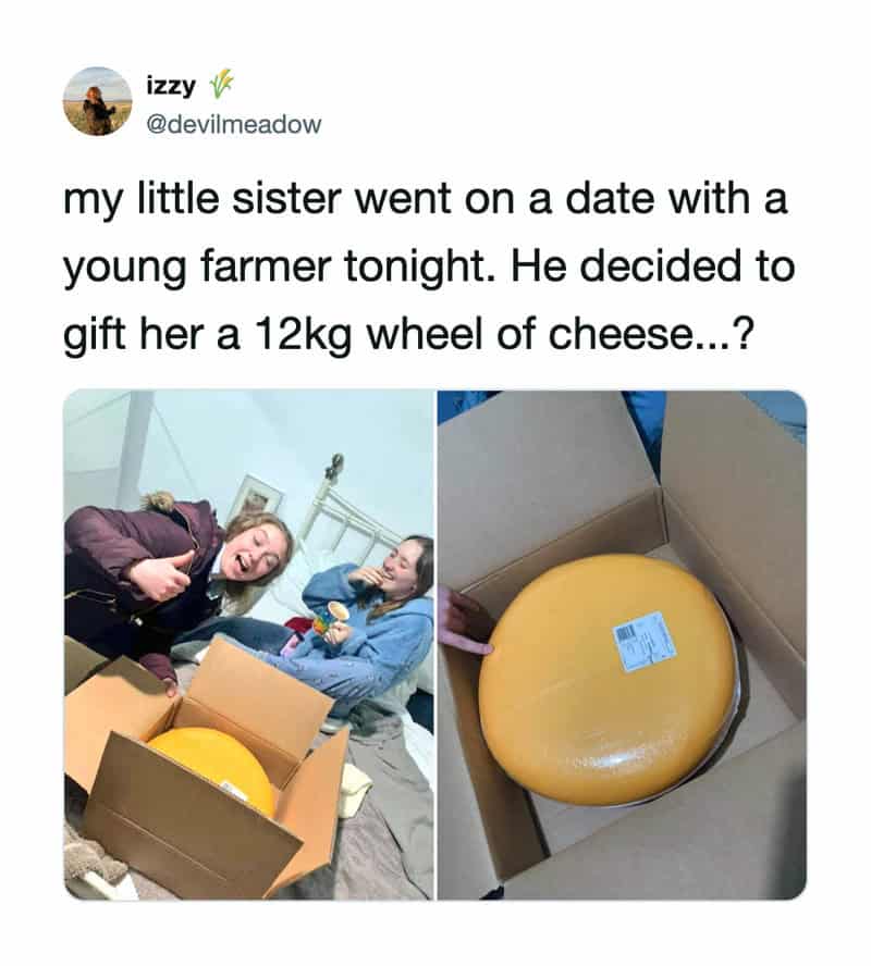 Two girls look surprised at a large wheel of cheese in a box.