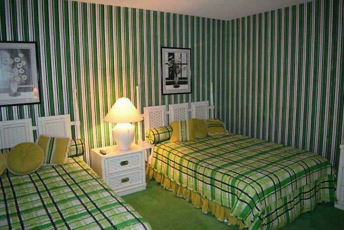 Bright bedroom with green striped wallpaper, two beds with plaid bedding, a white nightstand, and a.