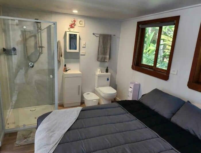 Small bathroom with shower, toilet, sink, and window in a cozy bedroom setting.