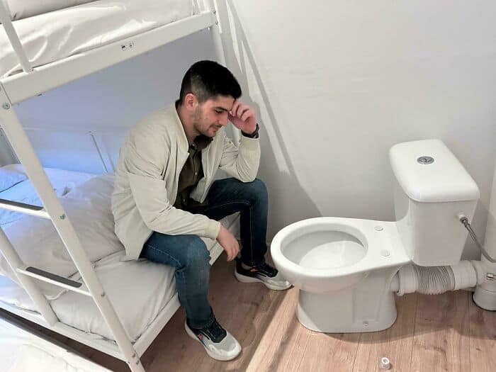 Man sitting on bed in a small room with a toilet nearby.