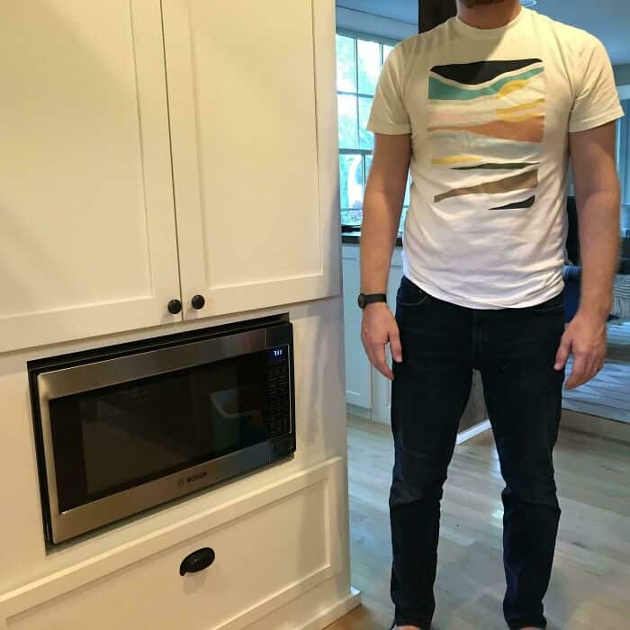Man standing in a kitchen with a microwave, wearing a colorful abstract t-shirt and dark jeans.