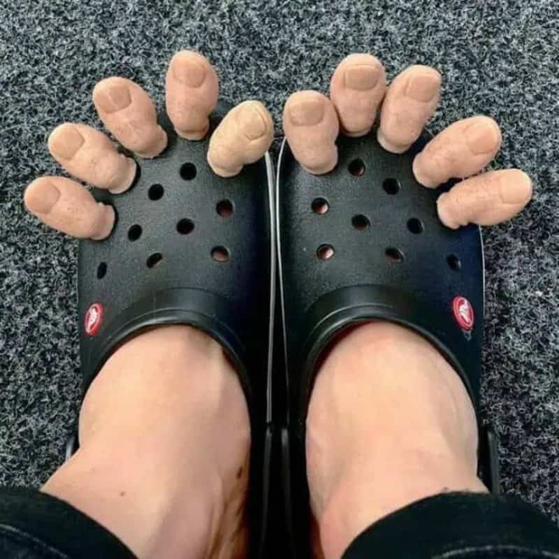 Worn black Crocs with fingers inserted into the holes, on a gray carpeted floor.