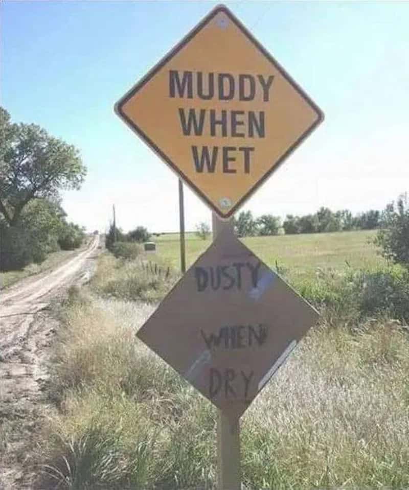 Muddy when wet, dusty when dry road sign in a rural area with grassy fields and a dirt path.