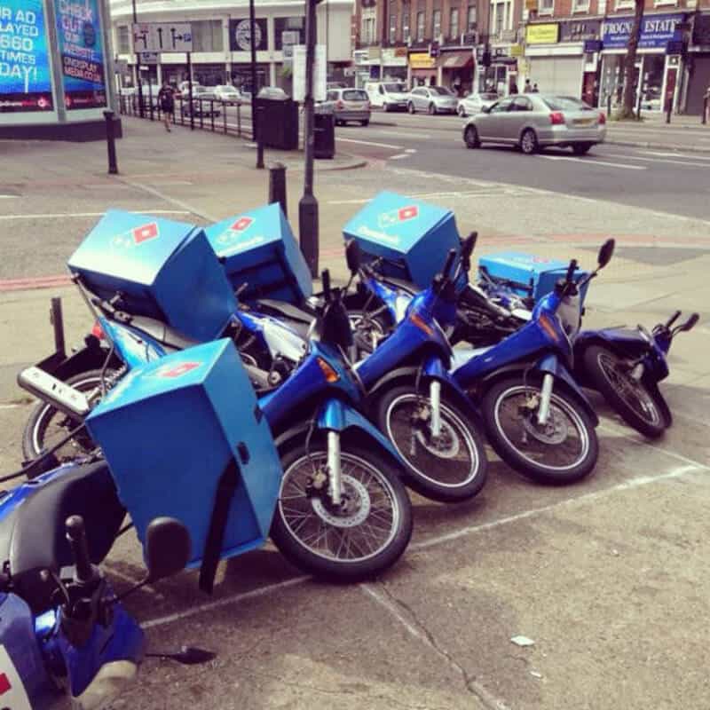 Four blue bikes with large blue delivery boxes parked on the sidewalk.