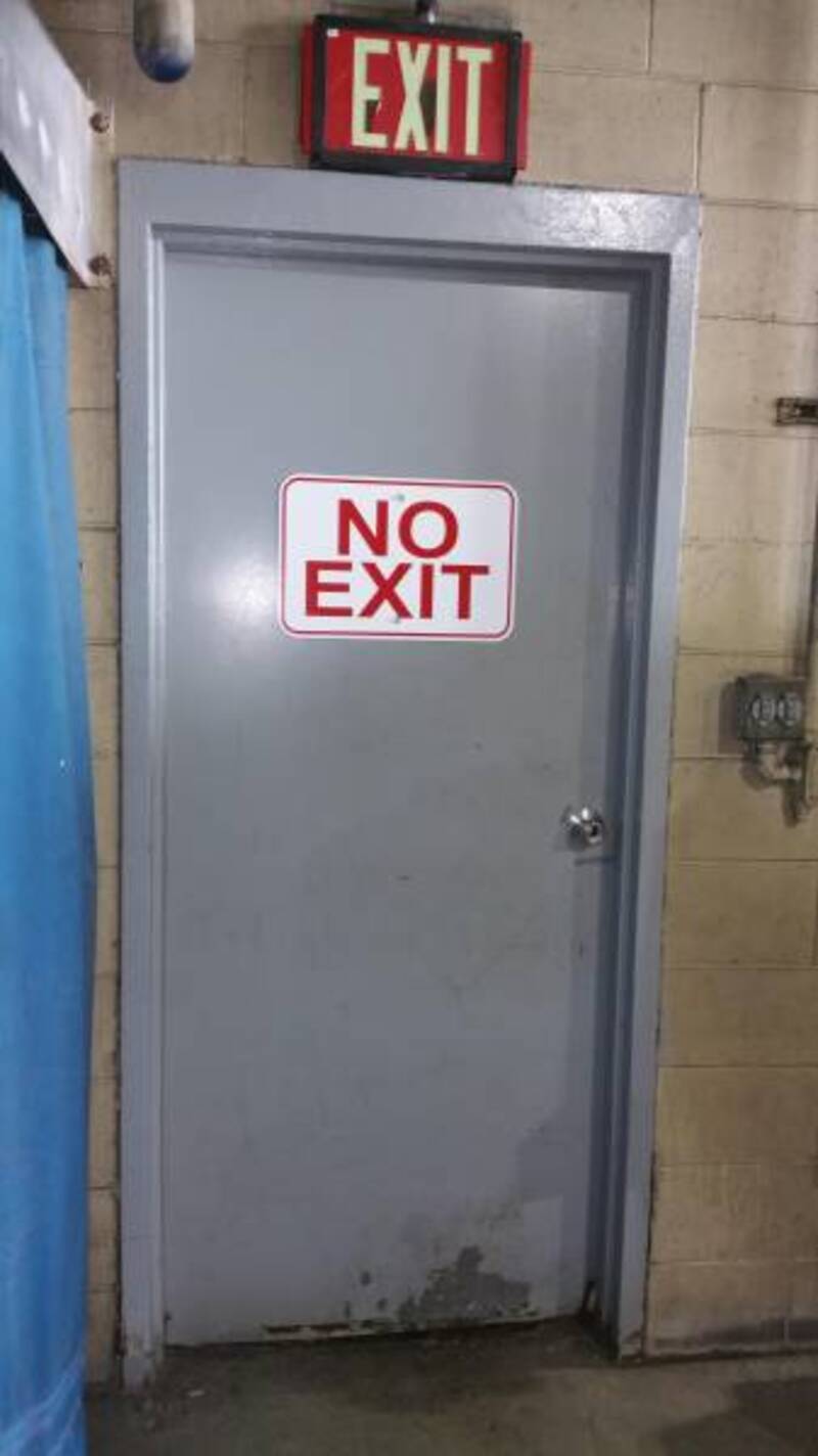 NO EXIT sign on a gray door with a "NO EXIT" sticker, located in an industrial or utility area.