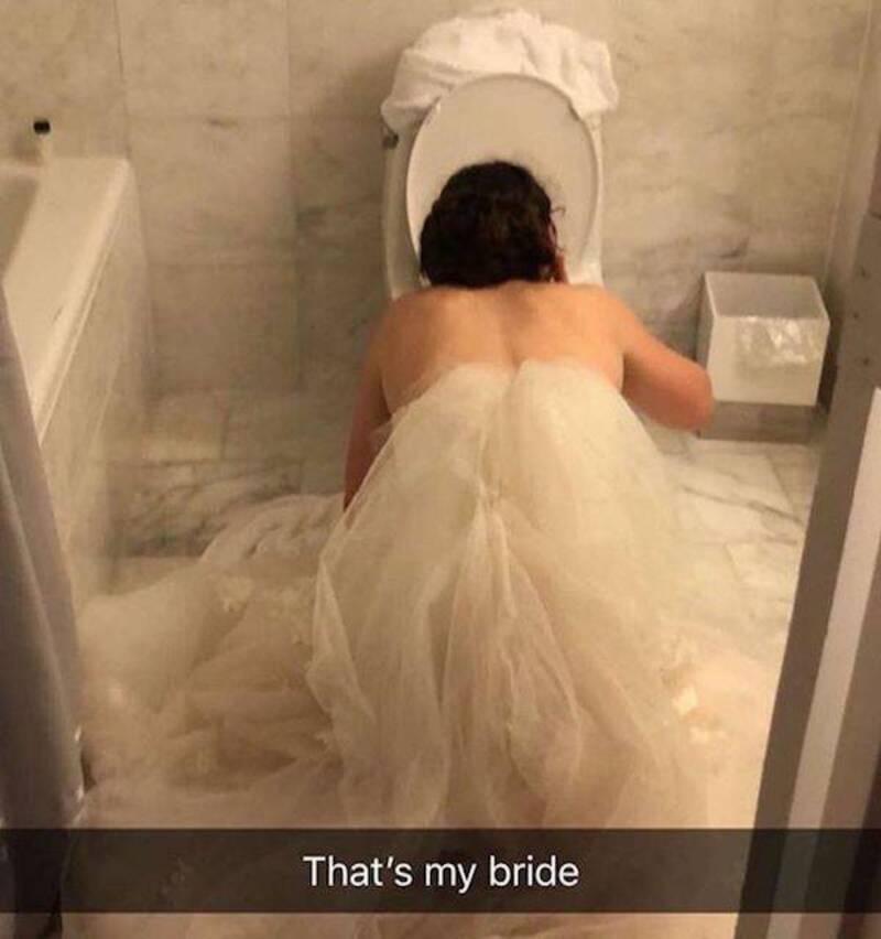 Bride in wedding dress leaning over toilet in bathroom.