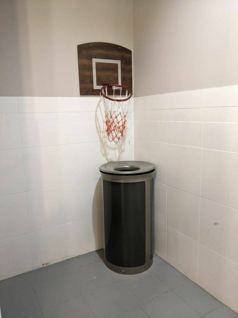 Basketball hoop attached to a trash can in a corner of a room with white tiled walls.