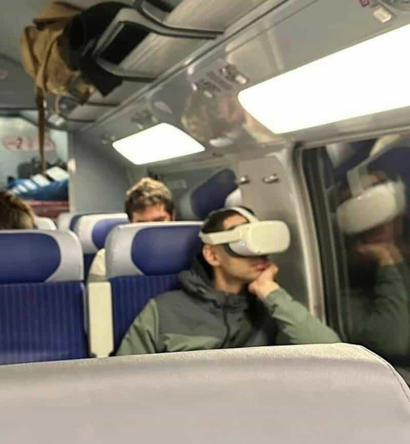 VR headset worn by a passenger on a train, sitting with hand on chin, other passengers in background.
