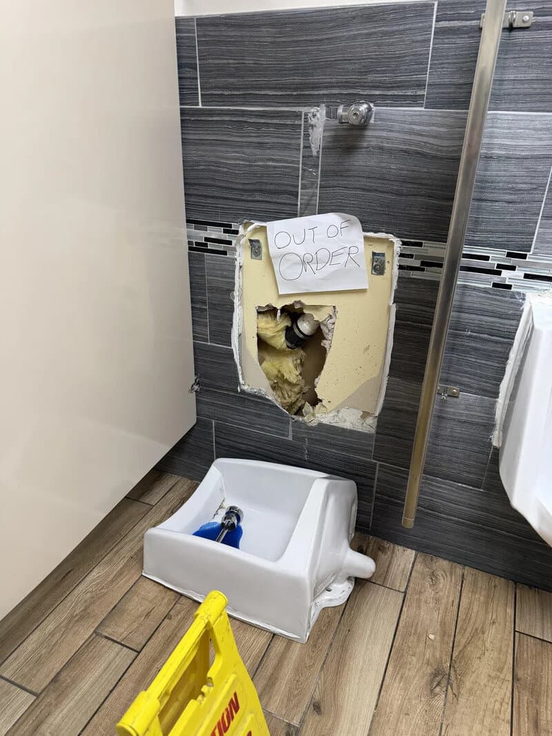 OUT OF ORDER sign on broken toilet with exposed plumbing and damaged wall, in a restroom.