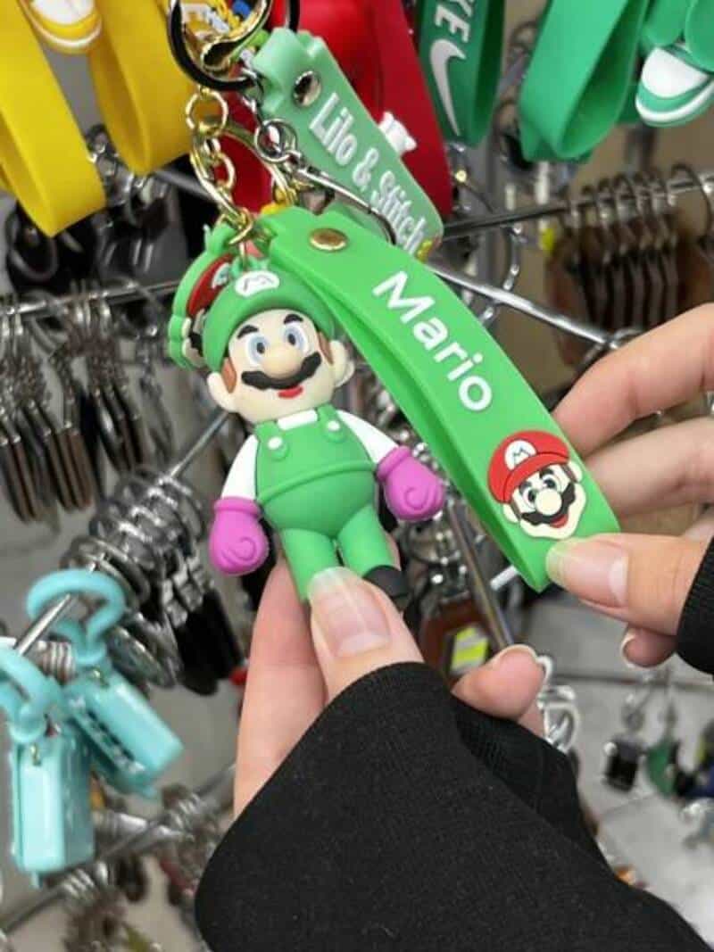 Mario keychain with green strap and Mario image, held by a person's hand in front of a keyring displ.