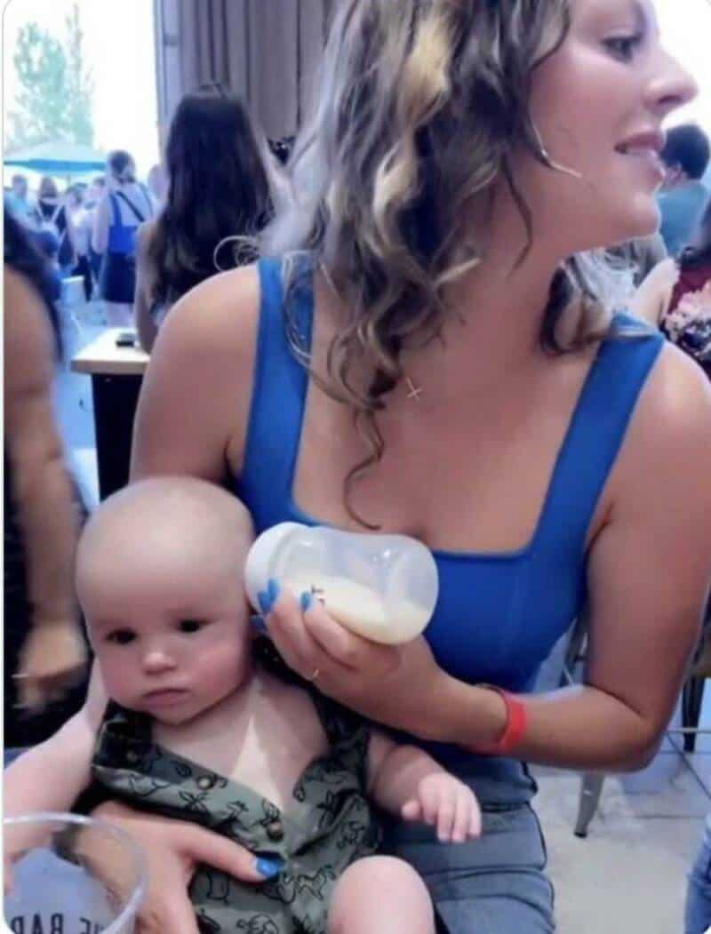 A woman holding a baby with a bottle in a busy indoor setting.