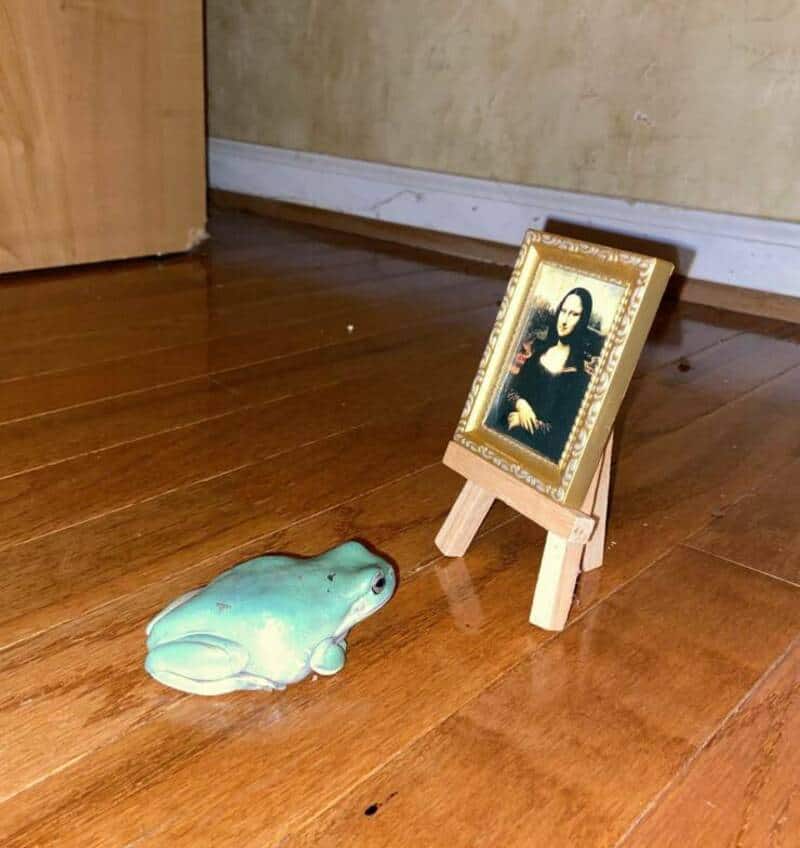 Small framed picture of Mona Lisa on a wooden easel and a ceramic frog figurine on a wooden floor.