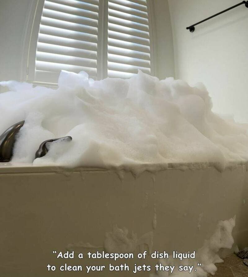 A woman relaxing in a bathtub filled with soap bubbles, near a window with white blinds.