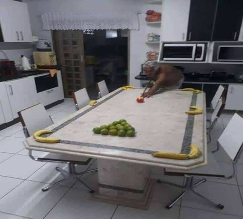 Man reaching for an orange on a kitchen table with bananas and green apples.