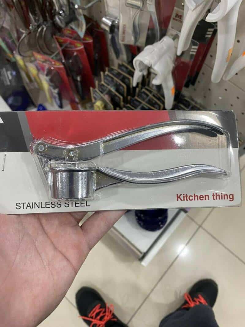Stainless steel garlic press kitchen tool in packaging, held in hand in store aisle.