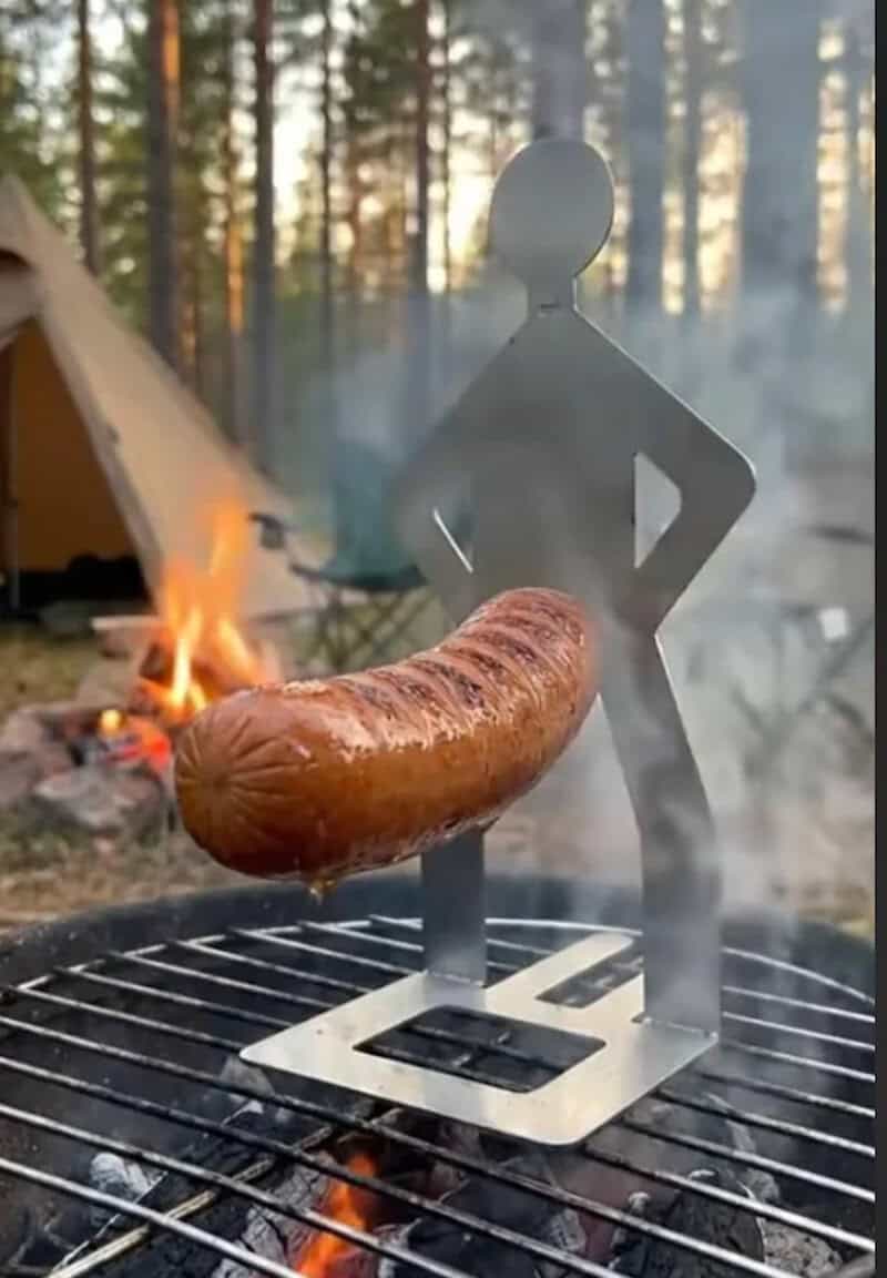 Metal figure holding a sausage over a campfire in the woods.
