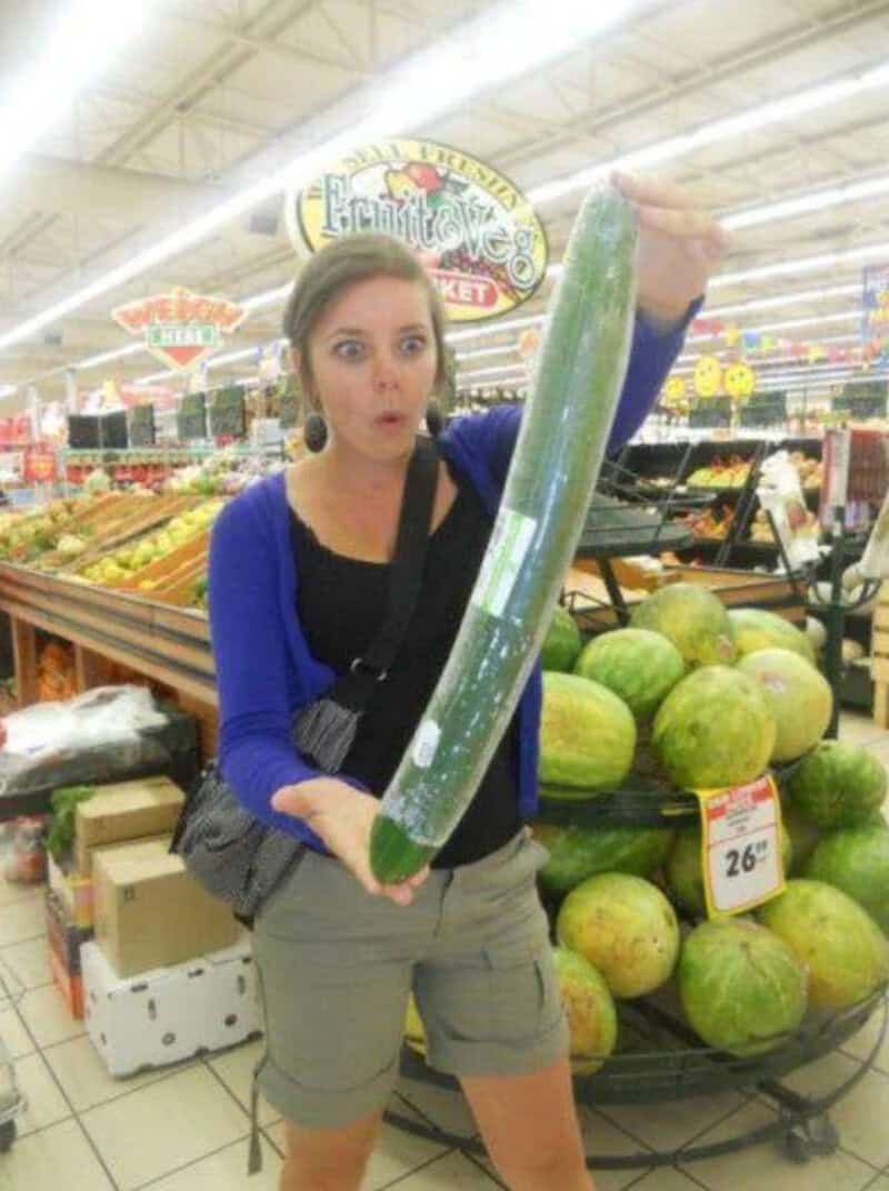 Woman holding a giant cucumber in a grocery store, surprised expression.