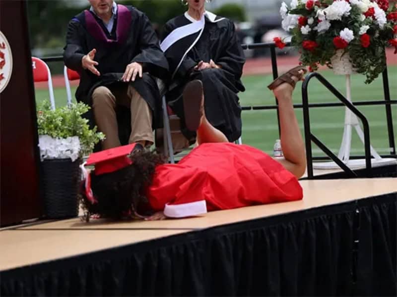 Graduate lying on stage with legs up, wearing a red gown and cap.