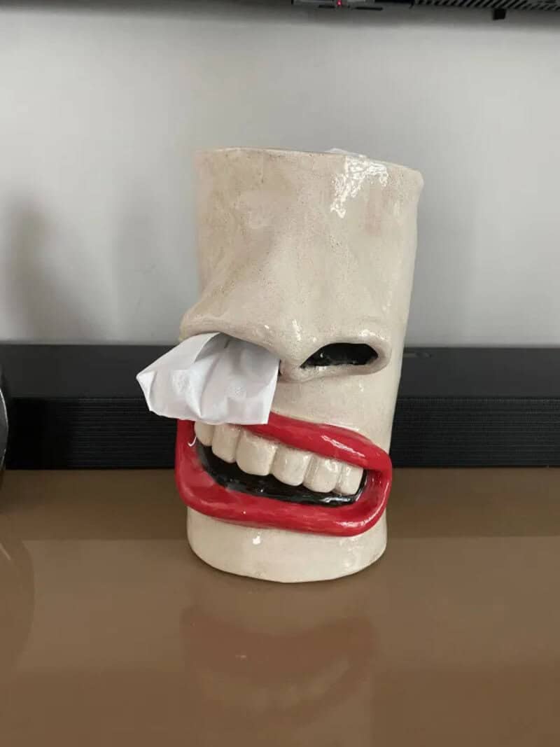 Ceramic mug with a face, tissue in nose, red lips, and teeth, on a table.