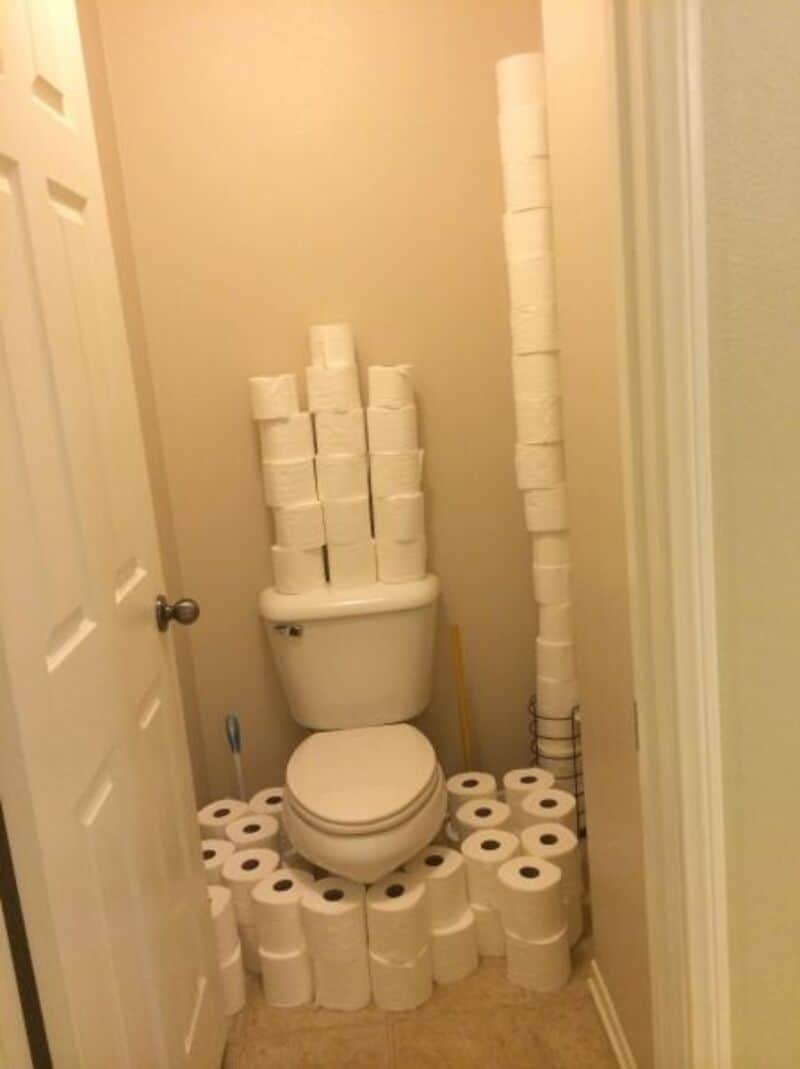 A toilet surrounded by stacks of toilet paper rolls on the floor, tank, and wall.