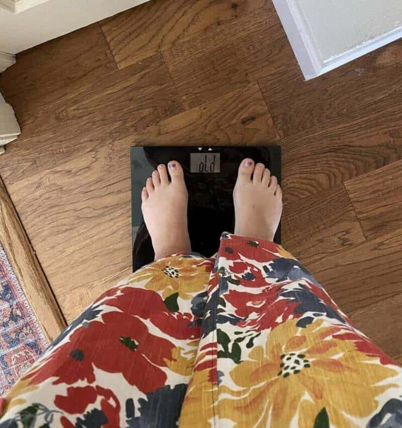 Person standing on a digital scale showing 100 lbs, wearing colorful floral pants.