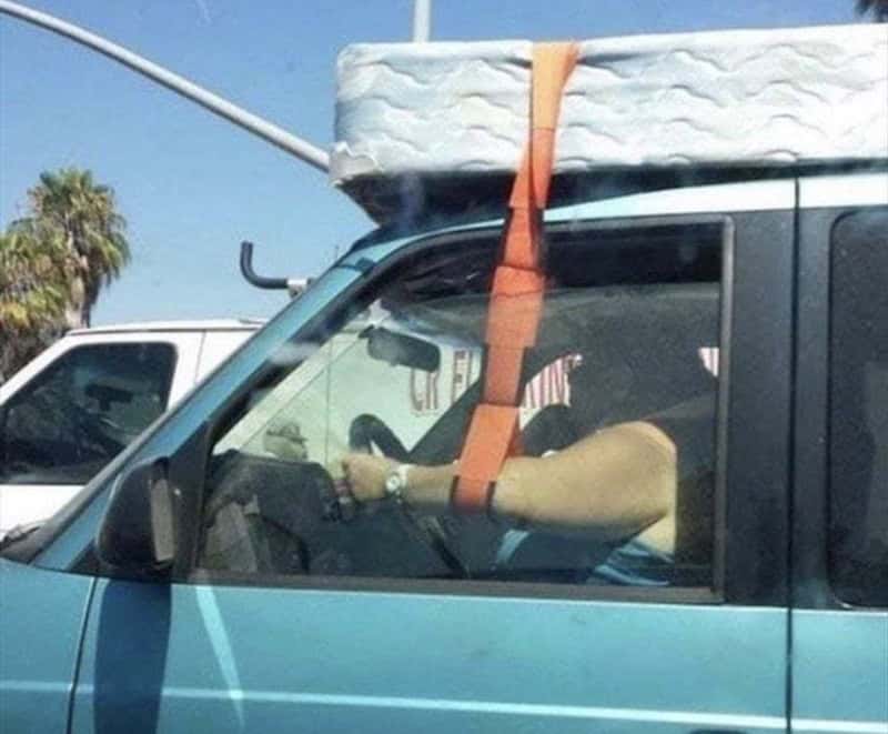 Person driving a blue van with a large object strapped to the roof.