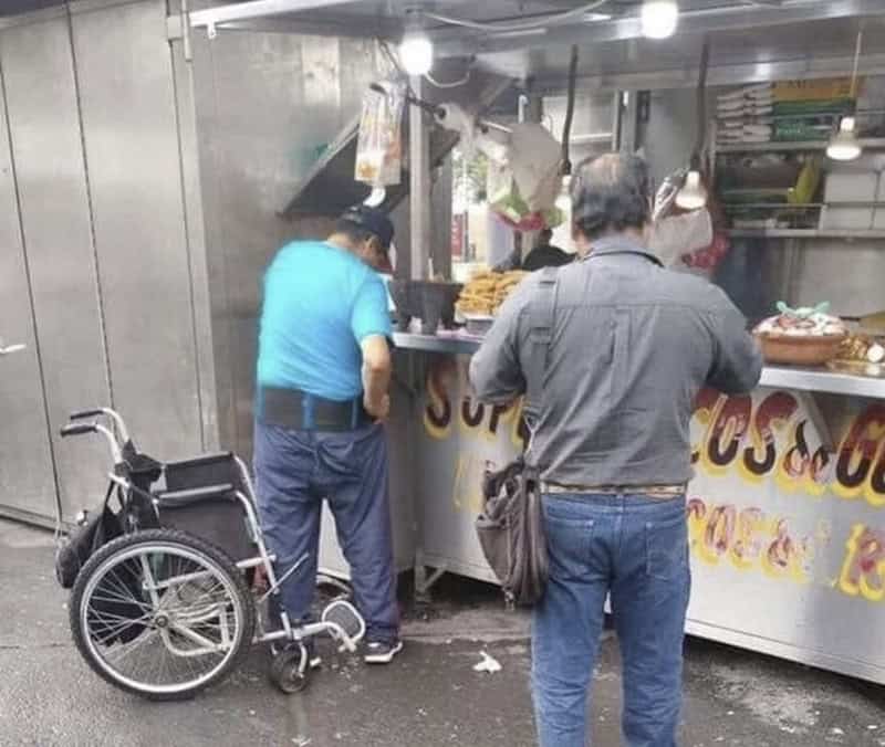 Two men at a street food stall, one with a wheelchair nearby, in a busy outdoor setting.