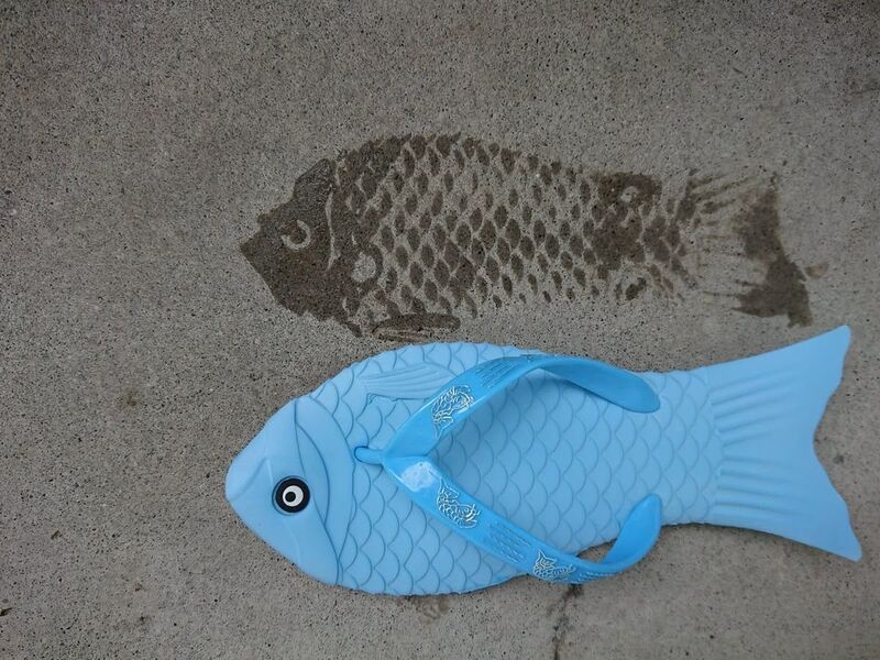 Blue fish-shaped kite on sand with a shadow of a real fish nearby.