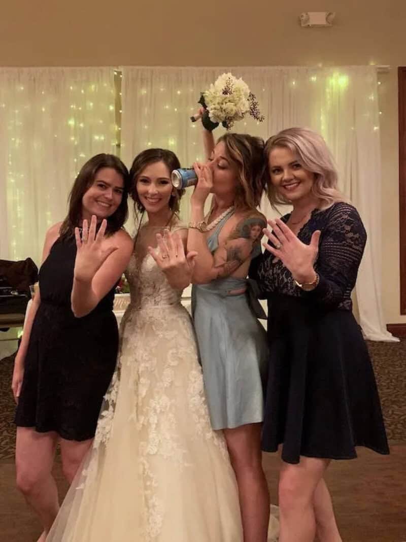 Four women at a celebration, one in a wedding dress, others in dresses, with one drinking from a can.