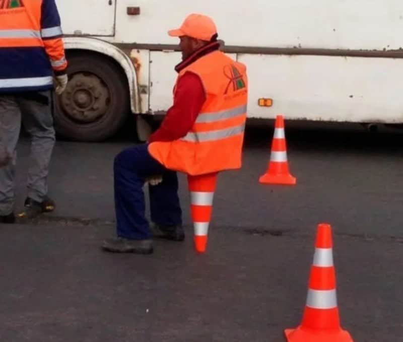 Person in orange vest sitting on a traffic cone, with other cones nearby.
