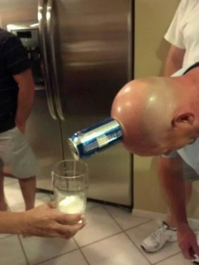 Man with a bald head holding a can in his mouth, pouring beer into a glass.
