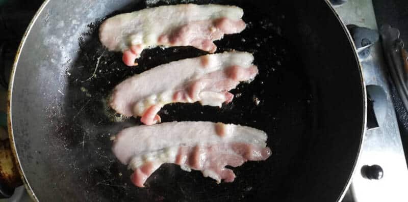 Bacon strips cooking in a frying pan.