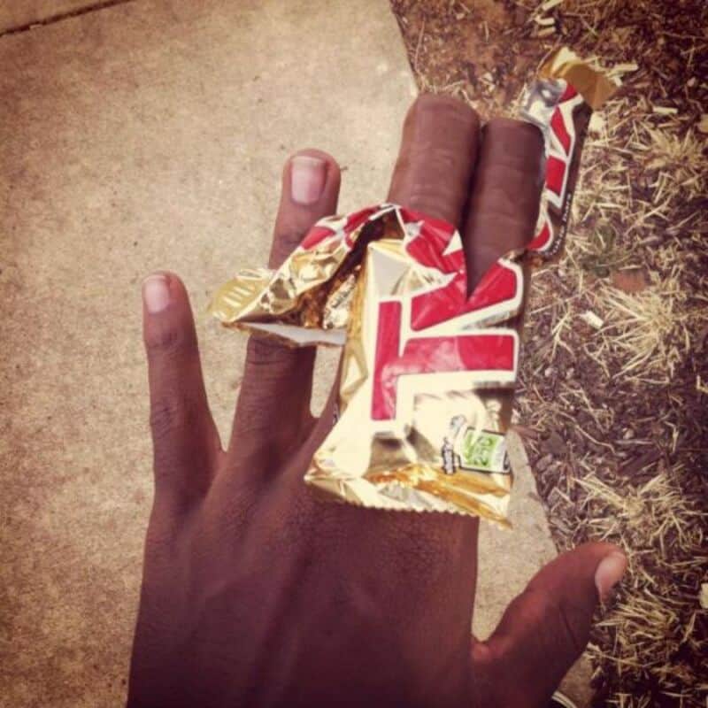 Hand holding a partially unwrapped chocolate bar with a background of grass and pavement.