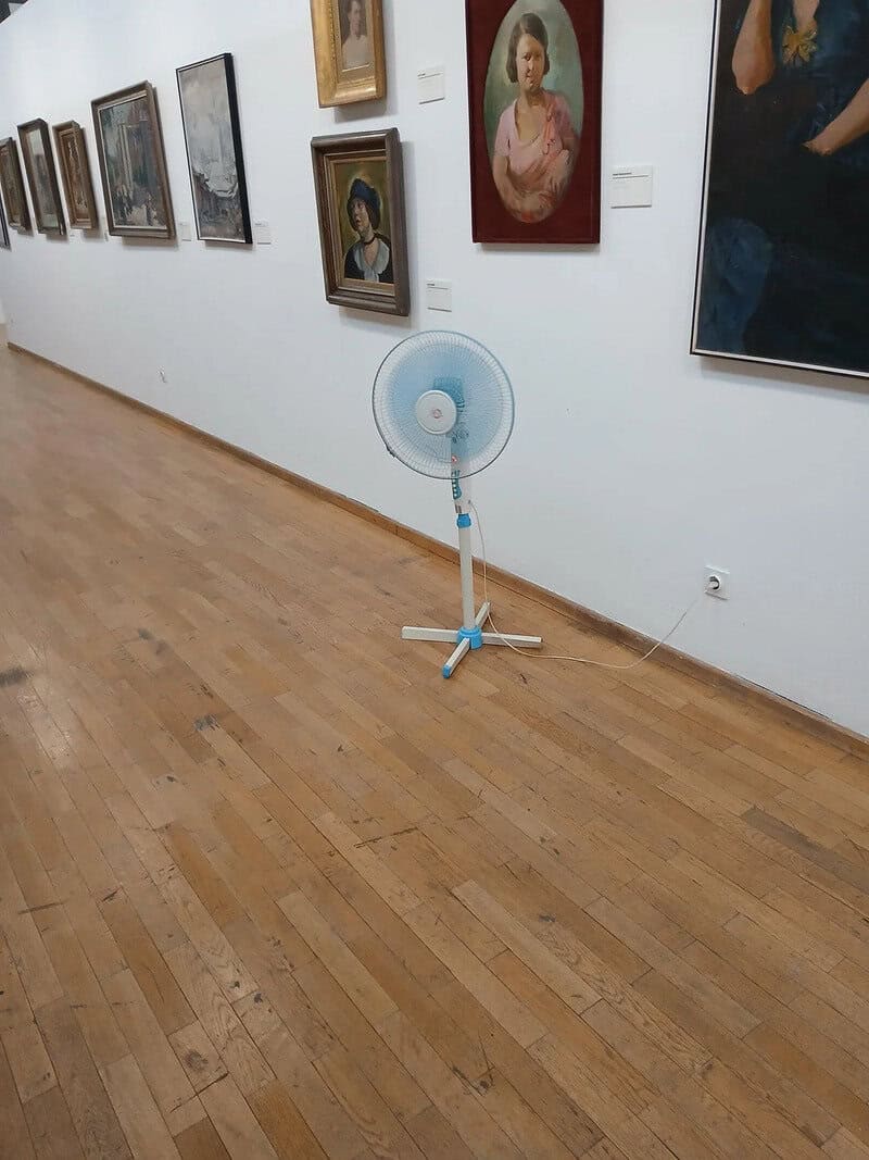 Fan placed in an art gallery with paintings on the wall behind it.
