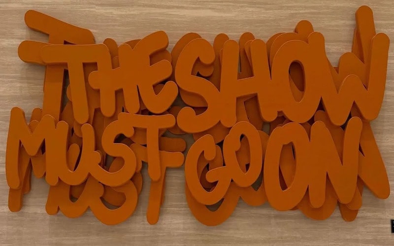 The show must go on! orange 3D letters on a wooden surface.