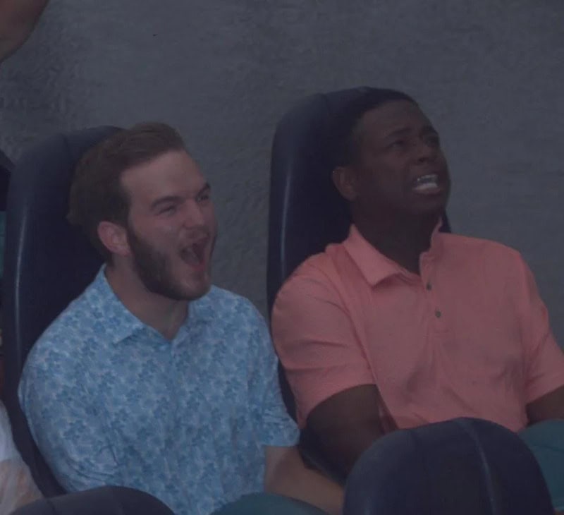 Two men sitting in a dark room, one laughing and the other looking distressed.