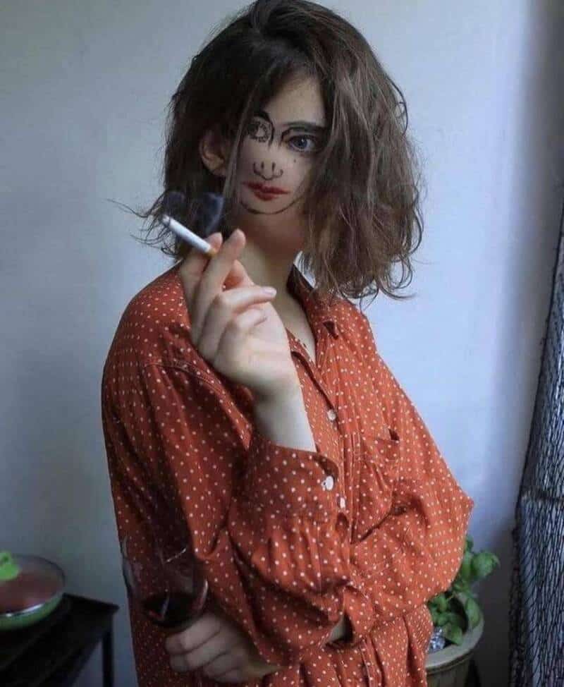 Young woman with messy hair, face painted like a clown, holding a cigarette.