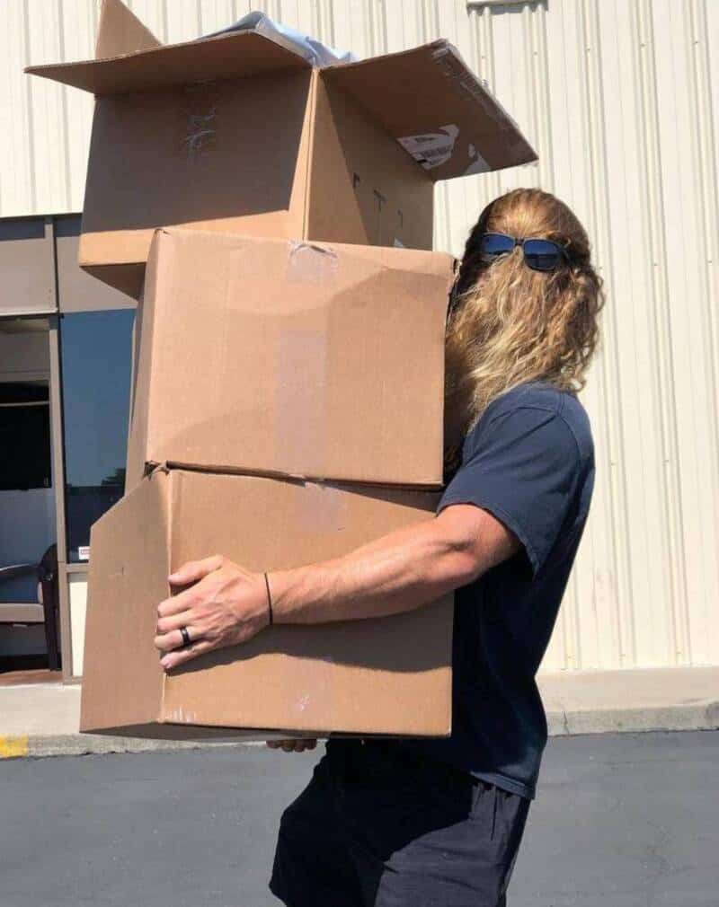 Person with long hair and sunglasses holding three cardboard boxes outside.
