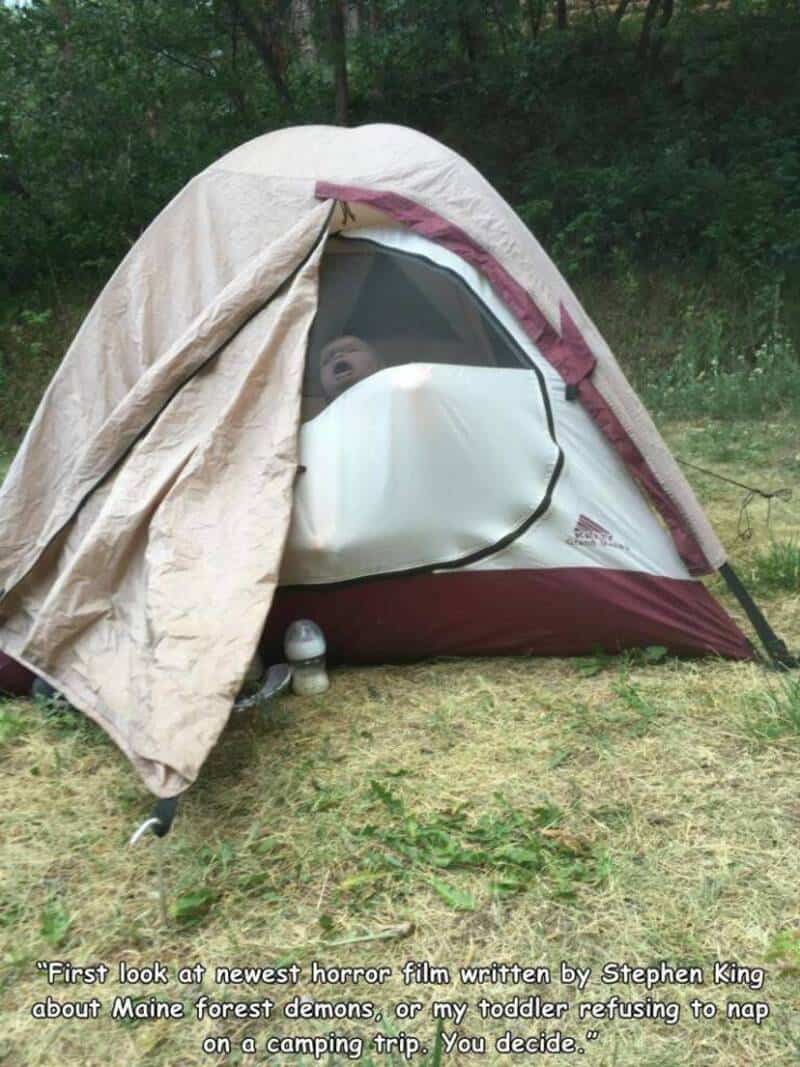 Child peeking out of a tent with a blanket draped over it, outdoors.