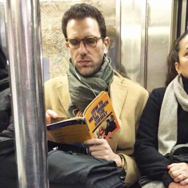 Man reading a book on a subway train, wearing glasses and a beige coat.