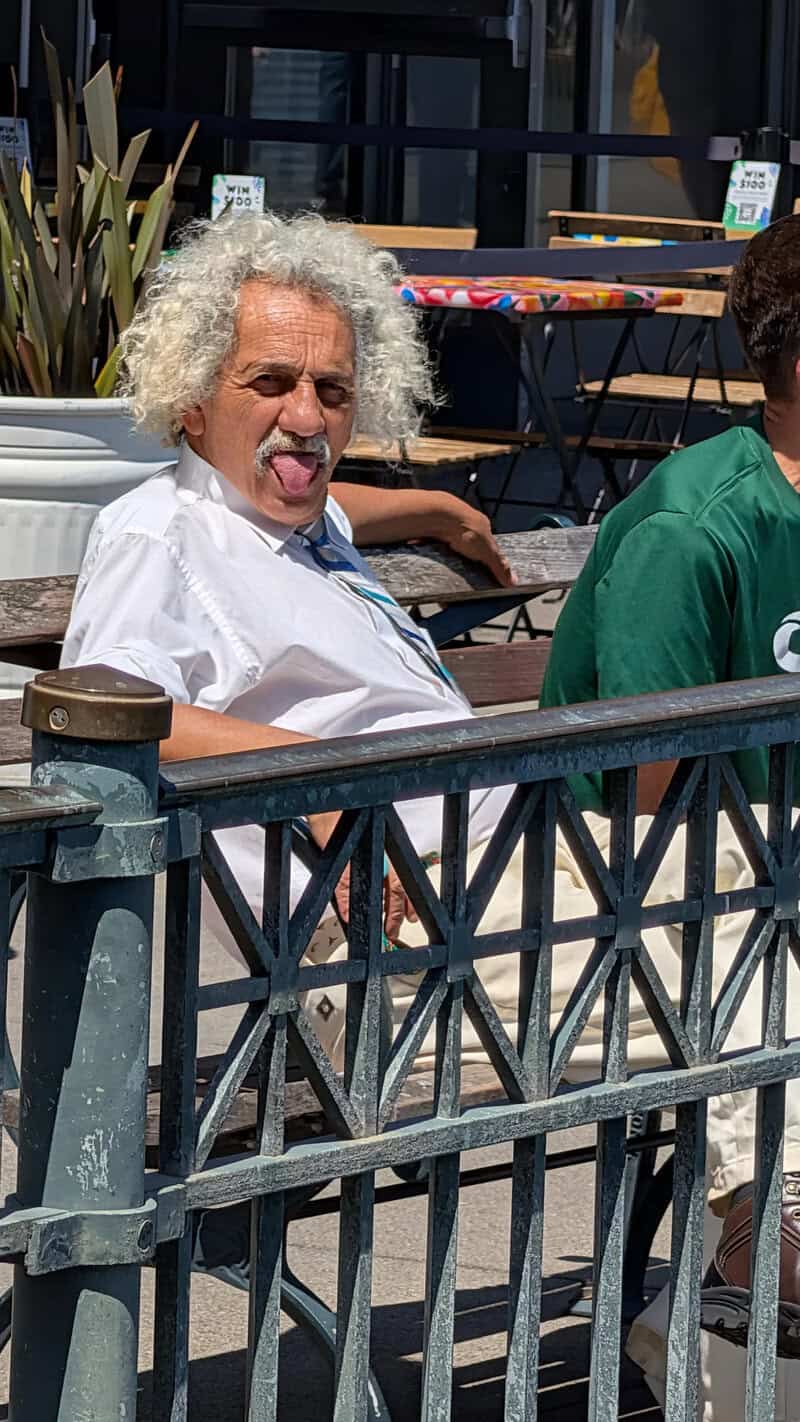 A woman with curly gray hair sticking out her tongue, sitting outdoors at a table.