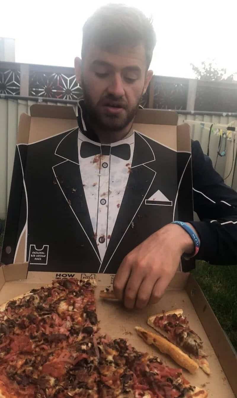A man with a cardboard cutout of a tuxedo on his chest, sitting outdoors with pizza in front of him.