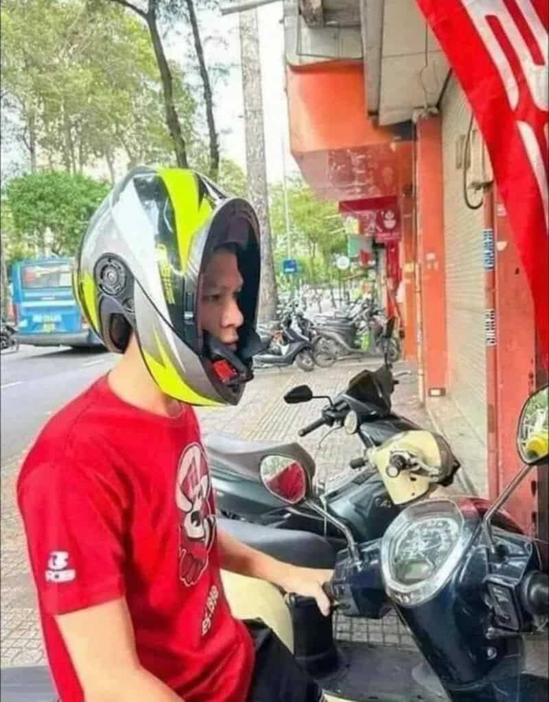 Young boy in a red shirt wearing a motorcycle helmet, standing next to a scooter.