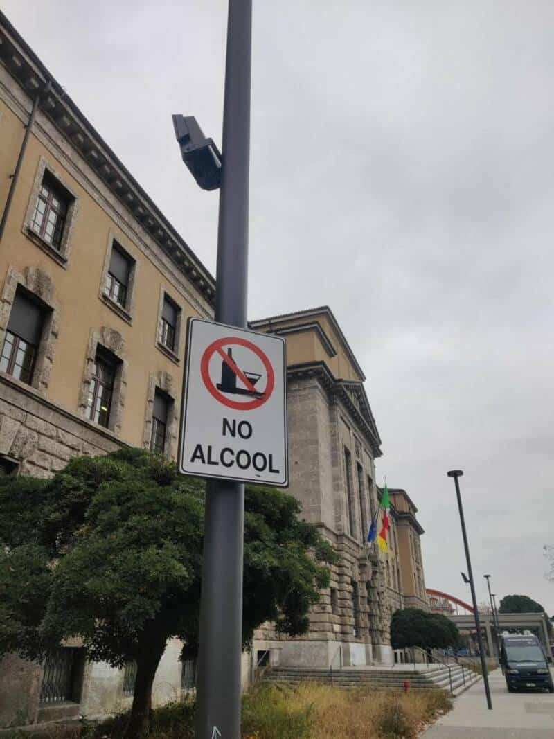 NO ALCOOL sign on a pole in front of a historic building with trees and a cloudy sky.