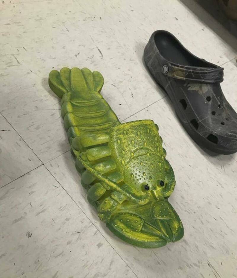 Green lobster-shaped slipper on a tiled floor with a black Croc shoe nearby.