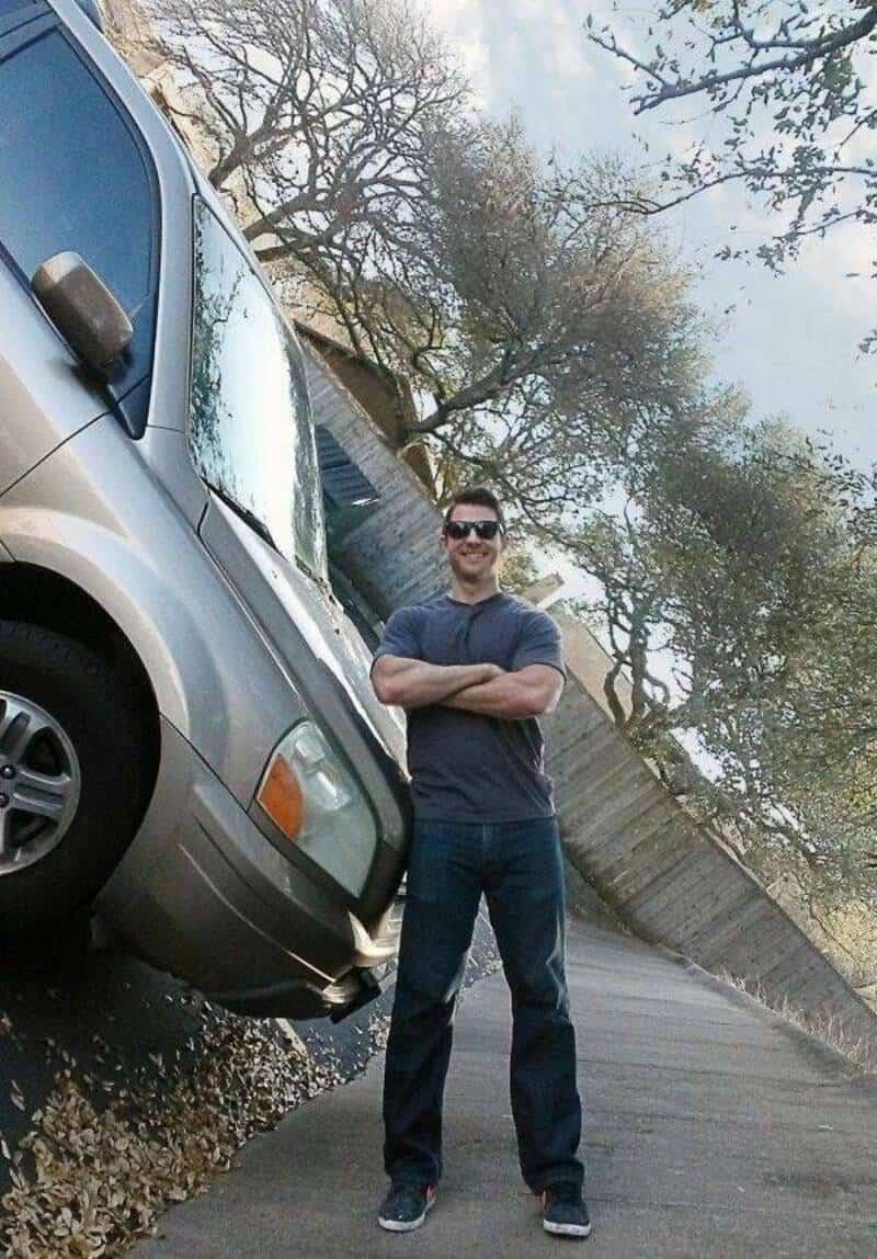 A man standing next to a tilted silver van on a sloped street.